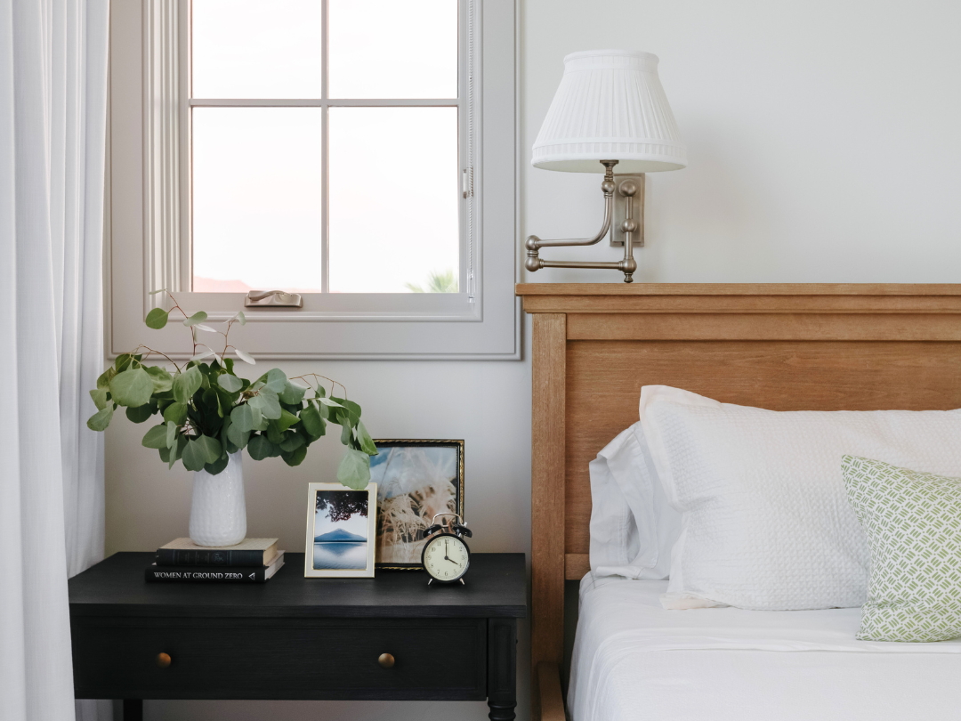 A close-up view of a bed and nightstand, featuring a few accessories, sconce and drapery.